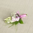 A delicate, handcrafted floral corsage or boutonniere rests on a softly textured, oatmeal-coloured linen background, the neutral fabric woven with fine threads that echo the understated elegance of a Queensbury wedding or prom. At the heart of the design is a compact cluster of soft, ruffled blooms in pale pink and blush tones, reminiscent of miniature stocks or spray carnations, each petal layered like a tiny cloud and gently fading at the edges. Between the pink flowers, airy sprigs of classic white baby's breath float like a light mist, their tiny blossoms adding a lacy, cloud-like texture. Threaded through the cluster are a few small florets of lavender or deep purple, quietly deepening the colour palette without overwhelming the soft romance of the piece. To one side, several creamy white to pale green buds, similar to freesia or slender lily buds, lie gracefully closed, suggesting flowers just on the verge of opening-perfect for a ceremony in Queensbury Park or a reception near the high street. The whole arrangement is cradled by lush, broad green leaves, their surface smooth and almost waxy, while fine, wispy tendrils of fern-like greenery reach out from beneath, giving a natural, slightly wild edge. The stems are neatly bundled and wrapped in a striking fuchsia satin ribbon, tied into an elegant bow with neat loops and short trailing ends that catch the light with a gentle sheen. The lighting is soft and even, with no harsh shadows, accentuating the freshness and careful craftsmanship. This refined floral accessory feels intimate and personal, ideal for pinning to a lapel or wrist on a special Queensbury celebration, from school leavers' events to quiet, romantic civil ceremonies.