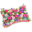 Seen from a gentle oblique, slightly elevated angle against a clean white backdrop, this pillow-shaped floral tribute reads like a tactile poem of colour and texture. At its heart, hot pink roses-some tightly furled, some just beginning to unfurl-create a velvety, resonant focal field whose passionate hue feels like a warm memory. Interspersed among them are soft, shell-pink spray carnations that introduce a delicate, ruffled counterpoint; their frilly edges catch the light differently to give the surface a layered softness. Here and there deep purple lisianthus lift their bell-shaped faces, adding cool contrast and a graceful, old-fashioned sentiment. Bright lemon-yellow ranunculus or small buttercup-like blooms appear as joyful punctuation marks, their layered petals catching a fresher, sunnier light. Architectural silver-blue eryngium thistles punctuate the composition with spiky, coastal texture, while abundant rounded eucalyptus leaves-silvery-green and cool to the eye-thread through the mass to provide breathing space and scent. The overall cushion rests upon a distinctly ruffled, deep red satin ribbon base; the pleated edging frames the flowers, reinforcing the rectangular pillow silhouette and lending a refined, ceremonial finish. The arrangement smells faintly of rose and eucalyptus, and its compact density suggests careful wiring and support beneath the blooms. Designed as a comforting sympathy or memorial cushion, it leans toward celebration of life rather than strict mourning, offering warmth, remembrance and quiet elegance. As a local composition often chosen for services near Queensbury station or small chapels along Kingsbury Road, it carries both floral craft and community tenderness in equal measure.