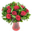 A lush, rounded bouquet of more than a dozen deep crimson roses fills this image with classic romance and polished design. Each rose is open and velvet-rich, the petals spiraling from a tight center into broad, sensuous curves that catch the light and suggest the soft, familiar scent of fresh blooms. The roses are nestled into abundant dark green foliage: flat, glossy leaves form a dense, verdant backdrop while broader emerald leaves have been artfully curled and rolled into sculptural loops, their smooth surfaces creating bright highlights and shadowed arcs that give the composition a modern architectural rhythm. The stems settle into a tall, translucent ruby-red glass vase whose subtle vertical ribbing twists faintly as it rises, catching reflected light in delicate lines that echo the roses' stems and reinforce the bouquet's rich palette of wine and leaf green. The arrangement sits against a crisp white background, isolating the intense reds and greens so the eye rests on texture - the velvet of petals, the glossy sheen of foliage, the cool, solid glass - and on feeling: an elegant declaration of love, deep warmth, and quiet luxury. As a local touch, imagine this bouquet prepared for a couples' anniversary delivery to a cosy flat near Queensbury tube station or placed on a dining table after a dinner on nearby Kingsbury Road; its refined presence works equally well as a centerpiece or a heartfelt gift. The lighting is soft and even, chosen to reveal each petal's detail and the vase's subtle refractions, making the whole image read as both intimate and timeless.