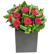 A deliberately modern bouquet captured in crisp light: deep, velvety crimson roses gather at the centre, each bloom opening in soft, generous spirals that catch the eye like small, sumptuous sculptures. Around them, broad emerald leaves have been folded and curled with practiced hands into overlapping loops and waves, their glossy surfaces catching highlights so the greenery reads as both texture and movement. The flowers sit low and confident in a matte black cube, a box-like vessel whose flat planes and soft finish provide a grounded counterpoint to the lushness above. The photographic lighting is bright and even, revealing the rose petals' satiny depth and the leaves' almost tropical sheen without washing out colour. Move in closer in imagination and you sense the contrast of textures: the silk-smooth petals against the firmer, waxy leaves, the slight give of petal edges and the clean, architectural line of the square vase. There is a subtle scent suggested by the image - classic rose perfume uplifted by green, almost citrus-bright notes from the foliage - a freshness that feels suited to an anniversary evening, a thoughtful romantic surprise, or to bring warmth to a modern living room or office. As a local florist who works around Queensbury and neighbouring Kingsbury and Kenton, I picture this arrangement gracing a café table near Queensbury station or arriving quietly at a front door, designed to make an intimate, memorable statement without fuss or frill.