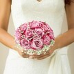 Imagine the anticipation of a wedding day in Queensbury distilled into a single image: a close-up of a bride's quietly excited grip on a radiant bouquet brimming with two-tone pink roses and airy clouds of white baby's breath. The roses, each petal brushing from soft, delicate blush to vivid magenta edges, seem to pulse with subtle energy, capturing the emotion of the morning. The bouquet is expertly shaped-round, dense, and artfully interspersed with gypsophila-so that every angle reveals new layers of texture and romance. The bride's hands, their tanness highlighted by a gentle natural glow, cradle the bouquet with tenderness, the light polish on her nails shimmering with each heartbeat. Her wedding dress, crafted from intricate white lace that flows into a gentle sash, provides a backdrop of classic elegance that mirrors Queensbury's blend of tradition and quiet charm. The softly muted background-the colour of warm sandstone-casts the bouquet and bride in an intimate light, focusing the gaze on their symbolic unity. This locally inspired scene speaks not just of beauty, but of the hopes and heartfelt joy behind every bouquet crafted for Queensbury couples beginning their new chapter.