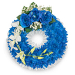 A carefully constructed circular wreath, its band formed from a dense, continuous ring of electric royal-blue chrysanthemums that read as a plush, almost velvet carpet of petals. The flowers are packed tightly so the surface appears luminous and saturated, each small bloom catching a cool, studio light that makes the blue feel vivid but calm. Around the inner and outer edges a fine fringe of tiny white florets - delicate sprays of baby's breath or similar fillers - softens the silhouette and provides a quiet, lace-like contrast to the saturated blue. Anchoring the composition at the very top are three full, velvety royal-blue roses, their petals unfurled with mathematical grace and gently tempered by small clusters of pure white gypsophila and a few deep green leaves that give the focal point depth and natural balance. From the left side a slender cascade of pristine white orchids, likely Phalaenopsis or Dendrobium, spills downward; their broad, open petals and elegant stems introduce a fragile, graceful movement. Intertwined with the orchids is a curved stem dotted with pale yellow-green buds, a subtle suggestion of new life and unfolding beauty. The wreath's inner rim is edged by a crisp, ruffled white satin or silk ribbon that frames the central void with a formal, clean finish. Prepared with the restraint and reverence appropriate for a memorial, the overall scent is faint and floral, the texture tactile, and the mood one of serene remembrance. Locally made in Queensbury with careful technique, this arrangement communicates peace, respect, and a quiet dignity suitable for funerals or memorial services.