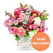 A romantic, garden-fresh bouquet fills the frame, its colours vivid against a clean white backdrop, as though waiting to be sent out for same-day delivery across Queensbury. At its heart are several large pink roses, some a deep, almost jewel-toned fuchsia with slightly darker outer petals, others a gentler, more muted pastel pink. These are embraced by plush carnations whose frilled petals range from warm coral and peach-pink to creamy white, creating a soft, inviting texture. Scattered among the larger flowers are petite daisy-like blooms in lavender and purple, their striking lime-green centres catching the eye and lifting the overall palette. The flowers are supported by a generous collar of foliage, where cool, silvery-green eucalyptus leaves mingle with richer, darker greens, giving the arrangement a natural, hand-gathered feel that would suit a home near Queensbury Circle or along Church Lane. A few reddish-brown leaves are tucked in like whispers of warmth, adding depth without changing the romantic character. All of these stems are carefully placed in a modern square clear glass vase, the transparent sides revealing the neat stems and fresh water. Around the lower part of the vase is a white band printed with tiny red hearts and the phrase all my love in looping red script, reinforcing the message of affection that the flowers themselves express. On the lower right of the image, a bold orange circular label reads VASE INCLUDED in crisp white capitals, drawing attention to the fact that the arrangement arrives complete and ready to display for birthdays, anniversaries, or thoughtful surprises throughout Queensbury.