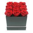 A luxurious square rose box arrangement fills this image, captured as if it were being presented by a Queensbury florist at a doorway on a quiet residential street near Mollison Way. The focal point is a dense cluster of twelve ruby red roses, all perfectly preserved and fully open so that every bloom reveals rows of velvety, overlapping petals swirling toward a tight centre. They are placed in three rows of four, packed neatly so their rounded forms touch and create a unified surface of deep crimson, with slight variations in tone hinting at the natural depth of the petals. The roses sit within a contemporary matte black square box, whose sharp edges and smooth finish provide a stark, elegant frame for the rich colour above. Around the lower section of the box runs a thin white stripe, a delicate decorative accent that cuts across the darkness and adds a refined, almost couture detail to the design. The entire arrangement stands alone against a pure white backdrop, without any other objects, so the eye is drawn immediately to the dramatic interplay of black, white, and red. Gentle, diffused lighting softens the scene, allowing subtle shadows to form between the petals and along the rim of the box, giving the roses a plush, almost tactile quality. This composition suggests an indulgent romantic gift, ideal for an anniversary surprise delivered across Queensbury, a discreet apology sent to a flat off Kenton Road, or a stylish piece of décor for a modern living space. It beautifully represents the trend for long-lasting preserved roses and high-end flower presentation in sleek, minimal hatboxes.