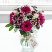 A vibrant mixed bouquet of fresh flowers stands proudly in a clear, textured glass vase, set against a softly glowing white background that feels like daylight streaming through a Queensbury window. At the heart of the arrangement are several large gerbera daisies in deep fuchsia and magenta, their velvety petals radiating out from dark, almost chocolate centres, immediately drawing the eye. Around them cluster bicolour chrysanthemums in rich purple and white, each petal tipped in plum tones that gradually fade to cream near the bright yellow centres, giving the bouquet a playful yet polished texture. Creamy white spray roses, some fully open and others still in tight, promising buds, nestle gently among the bolder blooms, adding a quiet elegance and a hint of soft fragrance. Sprigs of airy white gypsophila thread their way through the arrangement, like tiny clouds of blossom that lighten and soften the whole design. Deep green foliage frames the flowers, grounding the colours and giving the bouquet a natural, garden-fresh look, as if it had just been gathered from a well-kept home garden off Deansbury Drive. The squat jar-style vase is cinched at the neck with a rustic loop of twine, tied neatly in a bow, from which a small white gift tag hangs, decorated with a delicate pink rose illustration and subtle lettering. The tag suggests a personal message for a birthday, anniversary, or simple thank-you, the kind of thoughtful gesture often sent to homes and offices across Queensbury. The soft, diffused light highlights each petal, making the bouquet appear freshly arranged, full of life, and ready to brighten a dining table, bedside, or office desk with a sense of cheerful celebration and everyday joy.